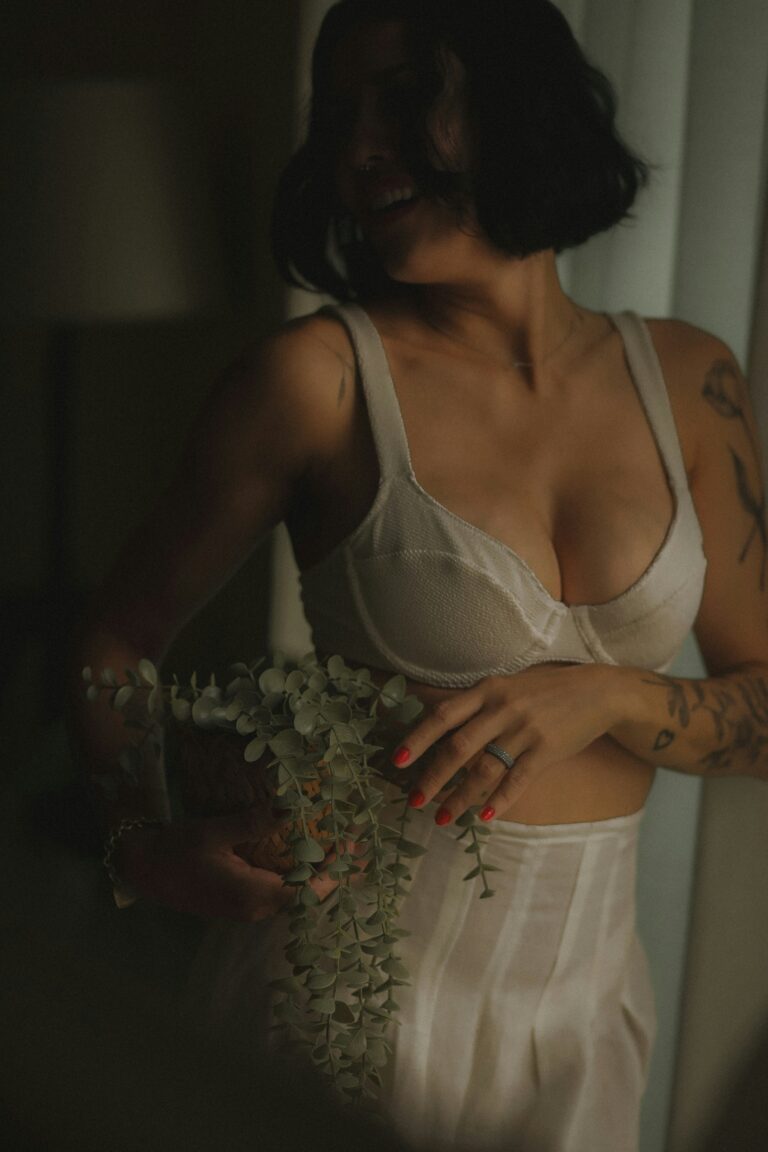 Tattooed woman in a white underwired bra holding a eucalyptus stem in soft moody natural light