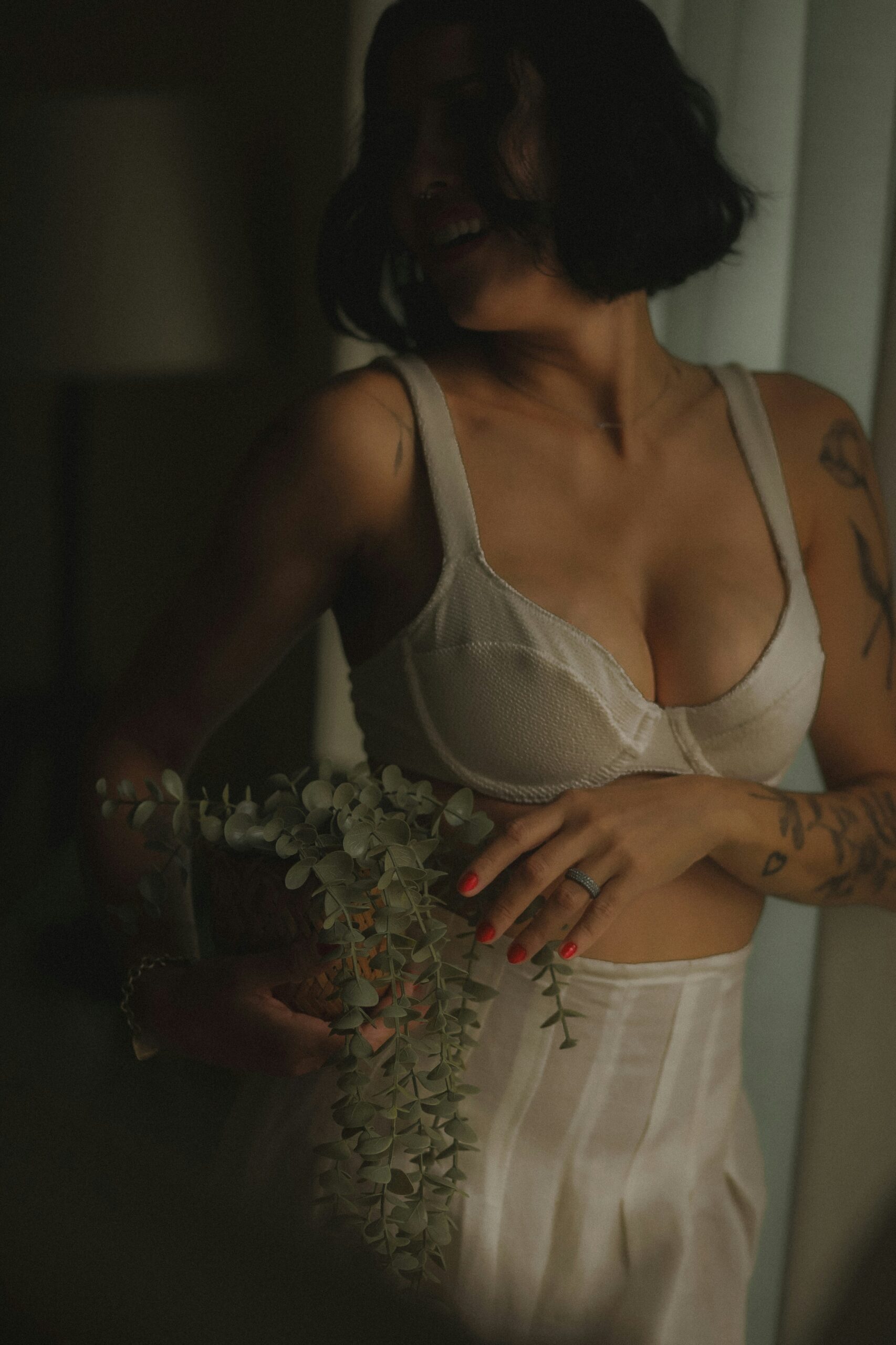 Tattooed woman in a white underwired bra holding a eucalyptus stem in soft moody natural light